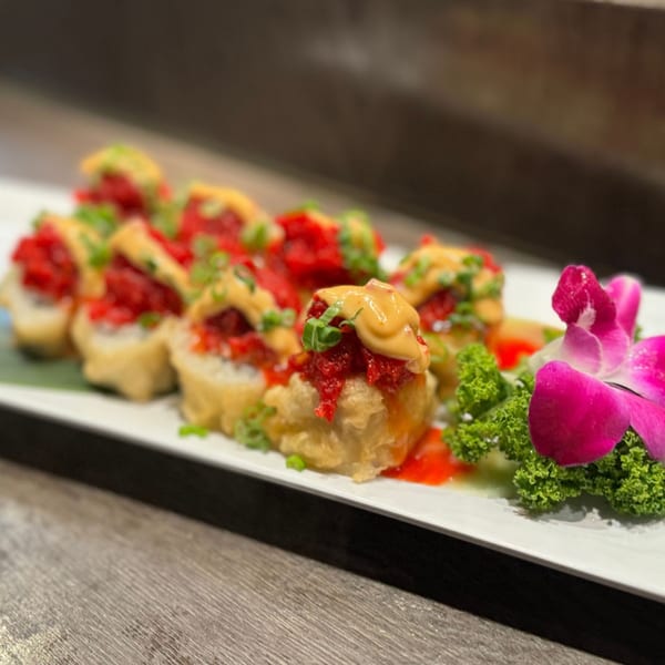 A plate of Koibito One Sushi & Teriyaki's deep-fried sushi rolls topped with red tuna, sauce, and green onions, garnished with a pink orchid.