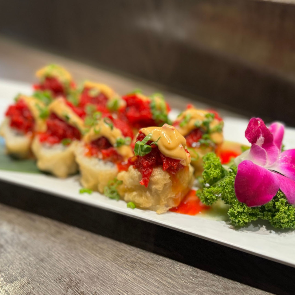 A plate of Koibito One Sushi & Teriyaki's deep-fried sushi rolls topped with red tuna, sauce, and green onions, garnished with a pink orchid.