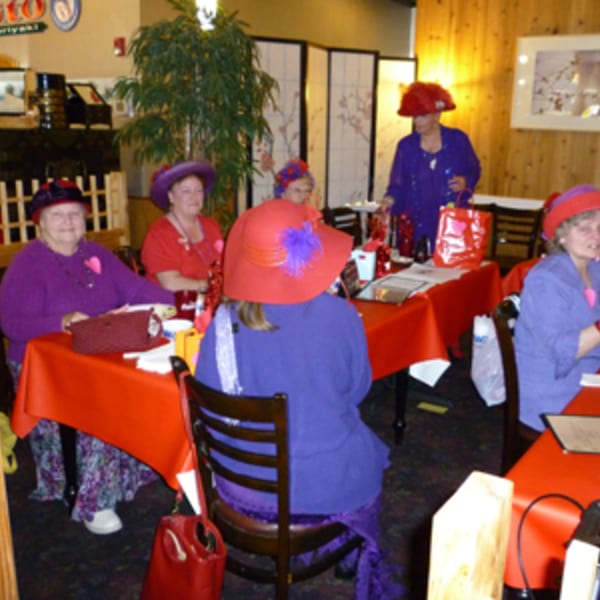 Women in red hats and purple outfits gather around tables with red tablecloths at Koibito One Sushi & Teriyaki.