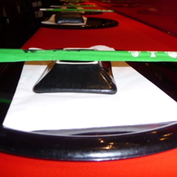 A row of place settings on a red table, each with a black plate, white napkin, and green chopsticks.