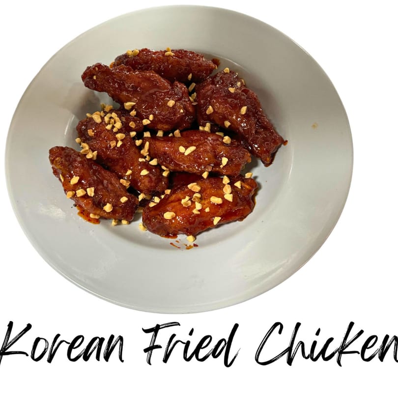 A white plate holds several pieces of Korean fried chicken, glazed and topped with chopped peanuts.