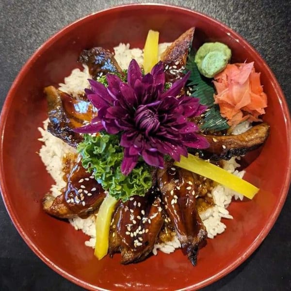 Unagi Donburi with grilled eel, rice, pickled ginger, wasabi, and a purple flower garnish in a red bowl.