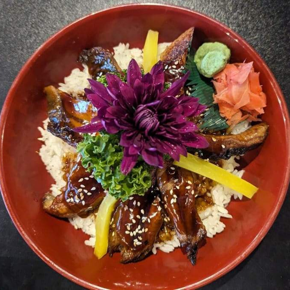 Unagi Donburi with grilled eel, rice, pickled ginger, wasabi, and a purple flower garnish in a red bowl.