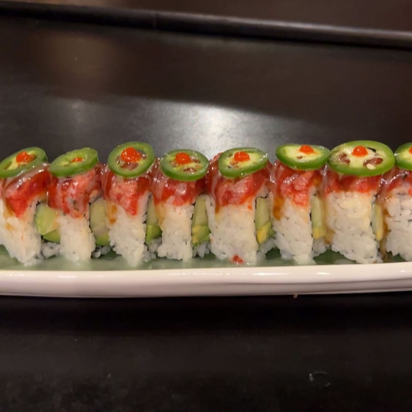 A row of Koibito One sushi rolls with avocado, red topping, and jalapeño slices on a white plate.