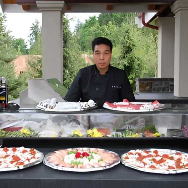 A Japanese chef stands behind a sushi bar with various sushi and sashimi platters.