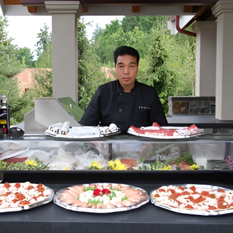 A Japanese chef stands behind a sushi bar with various sushi and sashimi platters.